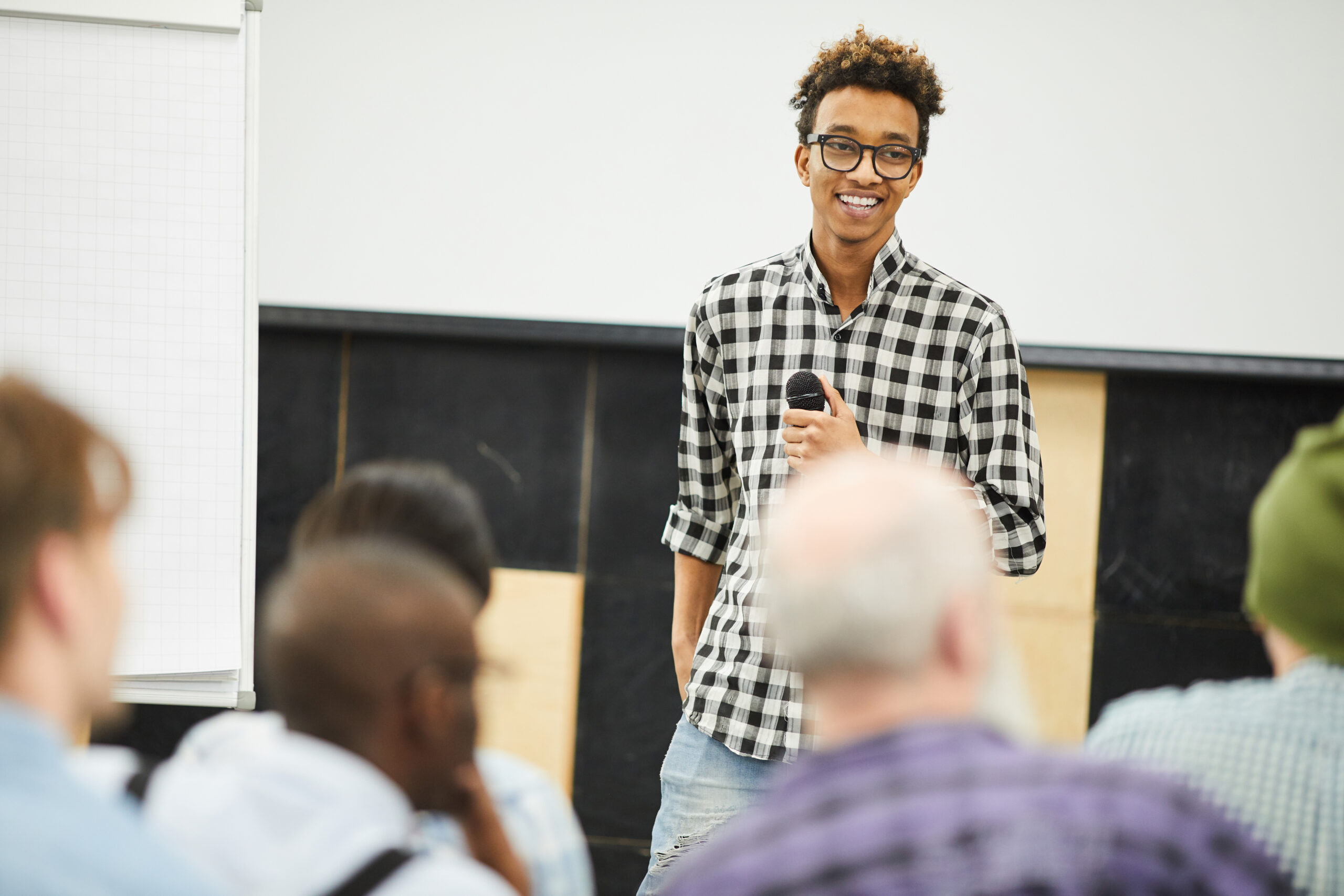 Positive successful young entrepreneur at business conference Positive successful young African-American entrepreneur in glasses standing in front of audience and laughing while talking to people at business conference