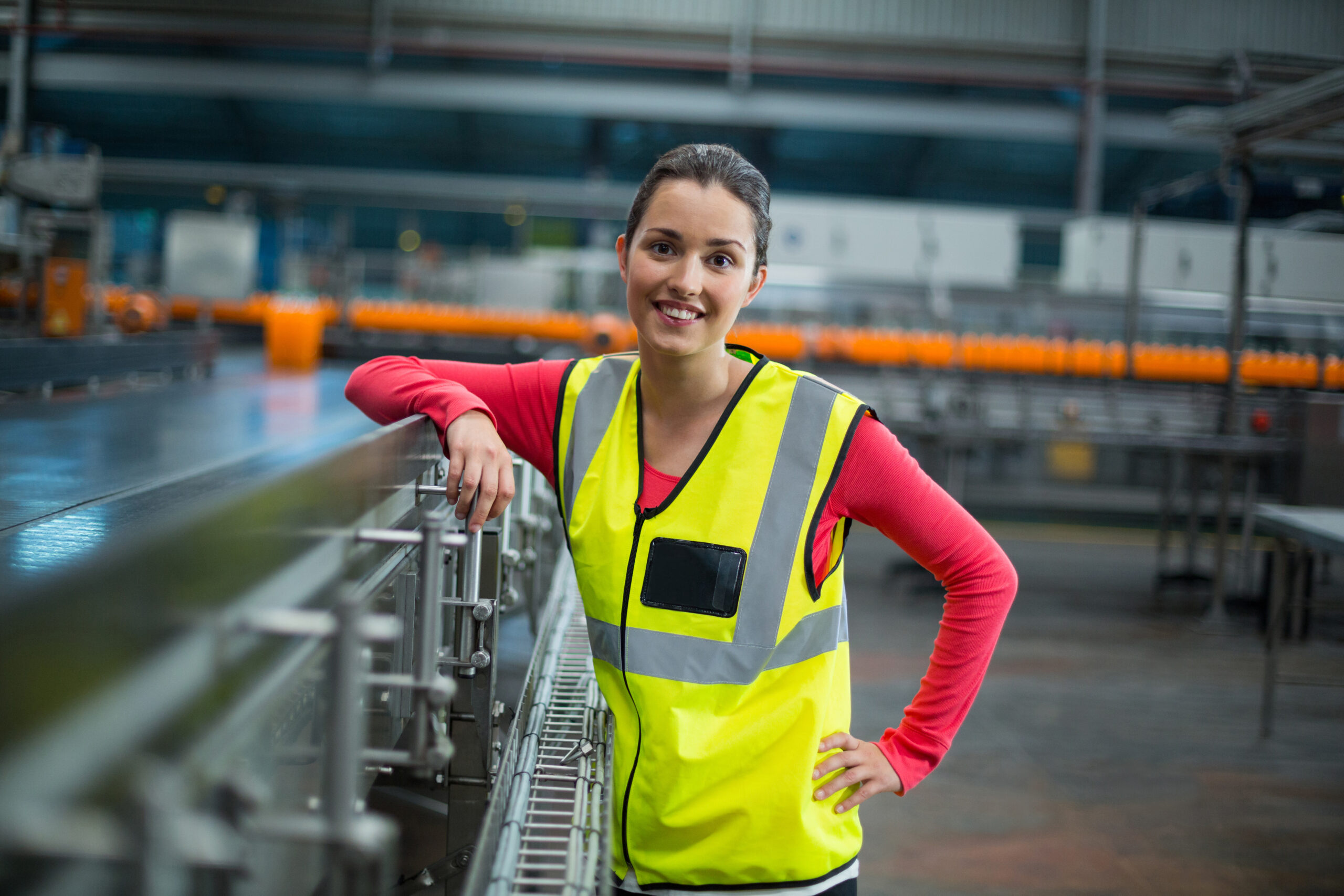 Smiling female factory worker standing next to production line Portrait of smiling female factory worker standing next to production line at drinks production factory